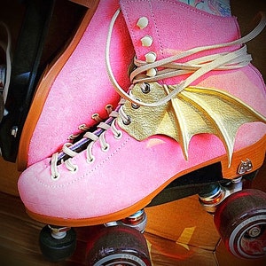 May include: Pink roller skates with gold bat wings attached to the laces. The skates have black wheels and a brown base.