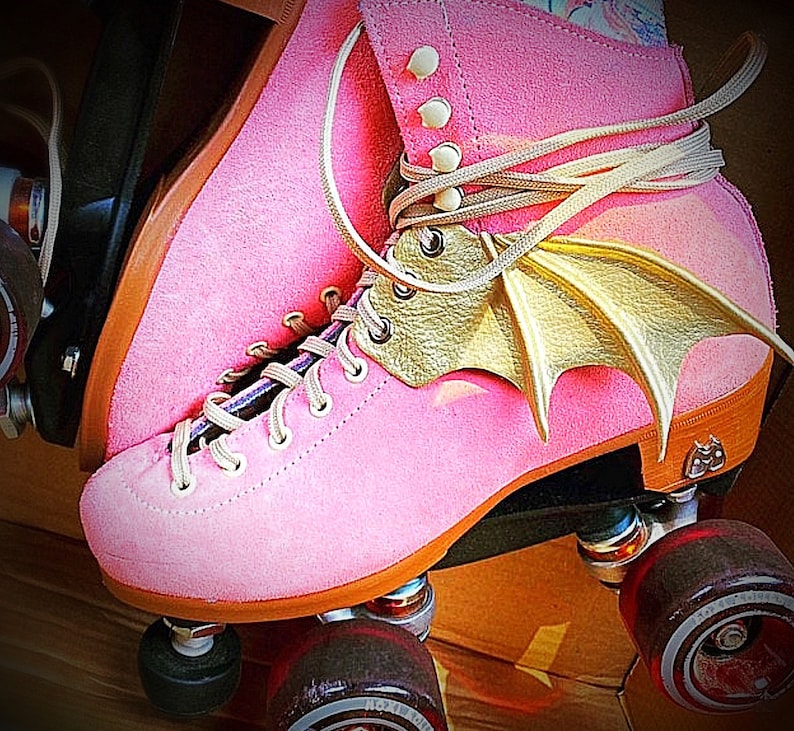 May include: Pink roller skates with gold bat wings attached to the laces. The skates have black wheels and a brown base.