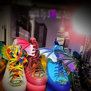 May include: Four pairs of high-top sneakers with colorful wings and laces. The sneakers are pink, blue, white, and black. The wings are made of fabric and are attached to the shoes with velcro. The laces are rainbow, purple, yellow, and black.