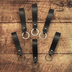 Może przedstawiać: Sześć czarnych skórzanych breloków do kluczy ze srebrnymi kółkami. Breloki są ułożone wachlarzowo na drewnianej powierzchni.