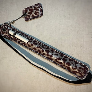 May include: A brown and silver leopard print pen case with a matching tag. The case has a black strap and holds a white pen. The tag is attached to the case with a silver chain. The case is designed to hold a pen.