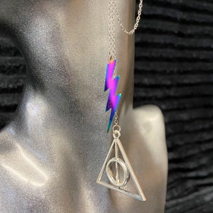 Op de afbeelding: Een zilveren kettingoorbel met een regenboogkleurige, iriserende bliksemschichtbedel en een zilveren bedel in de vorm van het symbool van de Doodsgeschenken uit de Harry Potter-serie.