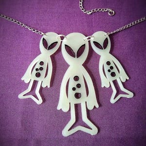 Könnte beinhalten: Eine silberne Kettenkette mit drei weißen Anhängern in Alienform. Die Aliens haben schwarze Augen und ein schwarzes Design auf ihren Körpern.