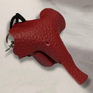 May include: A red leather keychain shaped like a saddle. The keychain has a textured surface and a small hole. A black cord and a silver keyring are attached to the top of the saddle.