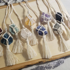 May include: A collection of various colored crystals wrapped in macrame with white tassels. The crystals are displayed on a wooden surface.