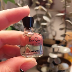 May include: A small, clear glass bottle of perfume with a black cap. The bottle is labeled "Parce Que Eau de Toilette" and "Capucci Paris".