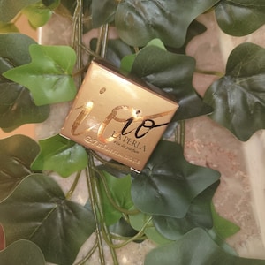 Puede incluir: Una pequeña caja dorada de perfume "Io La Perla", con letras doradas. La caja está rodeada de hojas verdes de hiedra. La caja contiene 7 ml de perfume.
