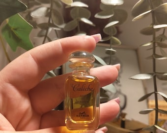 May include: A small, rectangular perfume bottle with a clear glass body and a rounded stopper. The bottle contains a golden-colored liquid and features the text "Calèche" and "Hermès Paris".