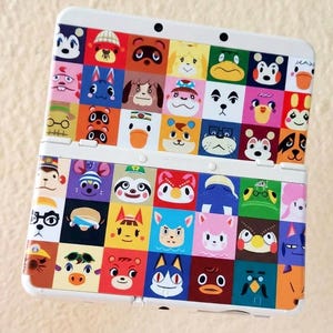 Puede incluir: Consola de juegos portátil Nintendo 3DS blanca con un diseño estampado colorido con varias caras de animales de dibujos animados. La consola está abierta, mostrando las pantallas y los botones.