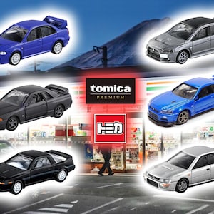 Może przedstawiać: Kolekcja sześciu miniaturowych samochodów z odlewu ciśnieniowego, w tym niebieskiego Mitsubishi Lancer Evolution, czarnego Nissana Skyline GT-R, srebrnego Subaru Impreza WRX STI, czarnego Toyoty Supry, niebieskiego Nissana Skyline GT-R i szarego Mitsubishi Lancer Evolution. Samochody są wystawione przed sklepem spożywczym z logo Tomica Premium.