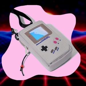May include: A light gray Game Boy-shaped crossbody bag with a black strap and zipper. The bag features a screen, buttons, and the text "Nintendo GAME BOY". The background is a pink shape with a retro-style design.