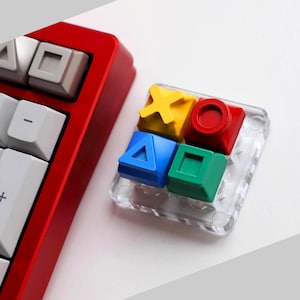 May include: A red mechanical keyboard with white and gray keycaps. A clear acrylic block holds four colorful keycaps: yellow X, red O, blue triangle, and green square. Gaming-themed.