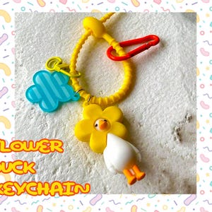 以下が含まれることがあります： 青い花のチャームと赤いクリップが付いた、黄色の花の鴨のキーホルダー。「FLOWER DUCK KEYCHAIN」の文字がオレンジと黄色で書かれています。