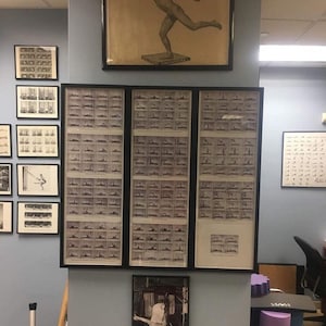May include: A wall with a collection of framed black and white photographs. The photographs are of people performing various exercises. The frames are arranged in a grid pattern. There is a poster of a man running on a platform with the text "SMALL UNIVERSAL" at the bottom.