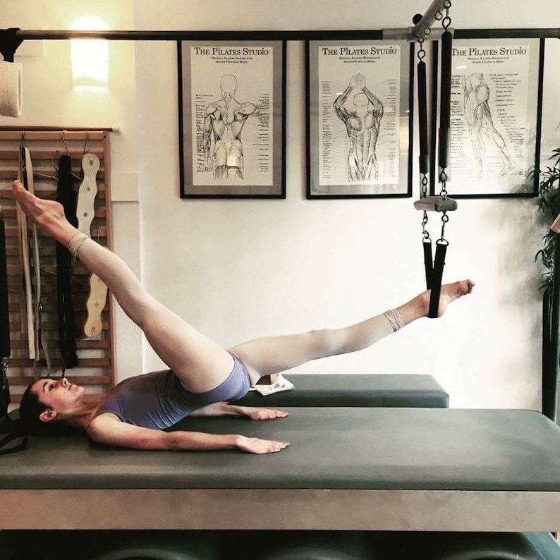 May include: A woman performs a Pilates exercise on a reformer machine. Her leg is extended and strapped to a cable system. The room has white walls and three framed anatomical charts with the text "The Pilates Studio".