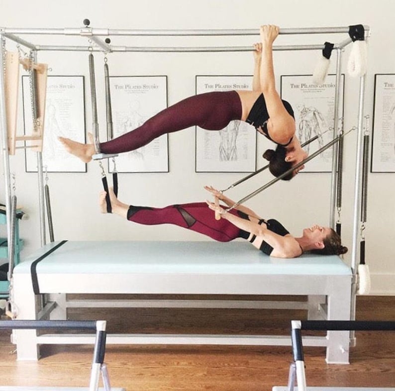 May include: Two people are doing a pilates exercise on a reformer machine. The person on top is holding onto the bar and is suspended in the air. The person on the bottom is lying on the reformer and is using their arms to push themselves up. The reformer is a piece of equipment that is used to strengthen and tone muscles.