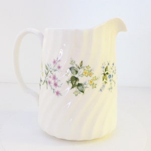 May include: White ceramic pitcher with a floral design featuring pink, yellow, and blue flowers. The pitcher has a twisted design and a handle.