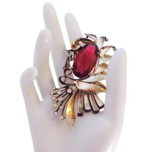 May include: Vintage brooch featuring a large, oval, ruby-red gemstone at the center. The brooch has a floral design with gold and silver-toned leaves and stems, accented with small, clear crystals. The brooch is set against a white background.