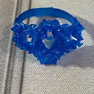 May include: A blue wax ring with a detailed, intricate design. The ring is made of a smooth, shiny material and has a delicate, feminine look.