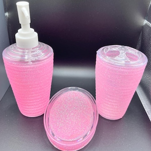 Op de afbeelding: Roze badkamerset met een dispenserfles, een tandenborstelhouder en een zeepbakje. Alle items zijn gemaakt van helder plastic met een roze glitterafwerking.
