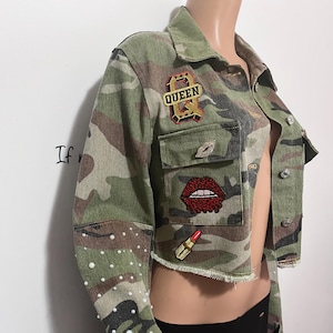 Puede incluir: Chaqueta corta con estampado de camuflaje con adornos de perlas en las mangas. Cuenta con parches que incluyen un lápiz labial rojo, un diseño de labios rojos y un parche "QUEEN". La chaqueta tiene un dobladillo deshilachado y botones.