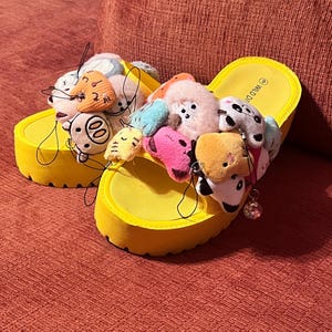 May include: A pair of bright yellow platform slide sandals featuring thick soles and textured footbeds. The wide straps are adorned with numerous small, colorful plush animal charms and character figures attached with thin black cords.