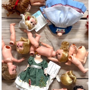 May include: A collection of vintage porcelain dolls with various outfits and hairstyles. The dolls are arranged on a wooden surface. Some dolls are missing their heads or limbs, creating a unique and whimsical display.