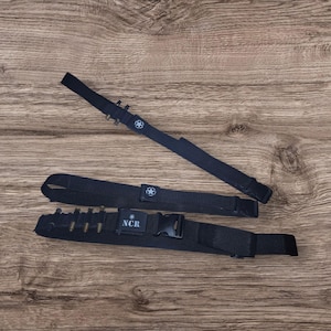 May include: Three black tactical belts with star emblems and a buckle. One belt has several bullet casings attached, and another has the letters "NCR" on a buckle. The belts are on a wooden surface.