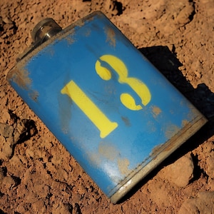May include: A blue metal flask with the number "18" painted on it in yellow. The flask is lying on a dirt surface.