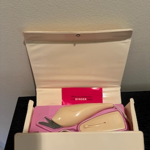 May include: A vintage Singer electric fabric scissors in a white case with a pink interior. The scissors are beige and have a cord. The case has the Singer logo on the front.