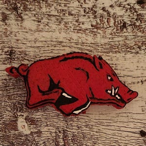 Pu&ograve; includere: Immagine stilizzata in rosso e nero di un Razorback, la mascotte dell'Universit&agrave; dell'Arkansas. Il cinghiale in corsa ha zanne affilate e un'espressione determinata. L'immagine &egrave; su una superficie di legno chiaro e strutturato.