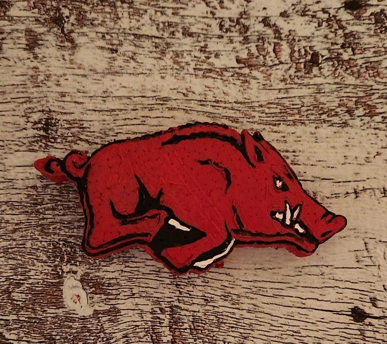 Pu&ograve; includere: Immagine stilizzata in rosso e nero di un Razorback, la mascotte dell'Universit&agrave; dell'Arkansas. Il cinghiale in corsa ha zanne affilate e un'espressione determinata. L'immagine &egrave; su una superficie di legno chiaro e strutturato.