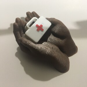 K&ouml;nnte beinhalten: Zwei braune, im 3D-Druck hergestellte H&auml;nde halten eine kleine wei&szlig;e Erste-Hilfe-Box mit einem roten Kreuz.