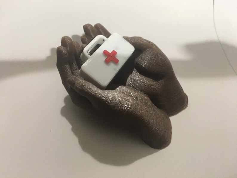K&ouml;nnte beinhalten: Zwei braune, im 3D-Druck hergestellte H&auml;nde halten eine kleine wei&szlig;e Erste-Hilfe-Box mit einem roten Kreuz.