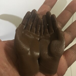 K&ouml;nnte beinhalten: Ein Paar braune, 3D-gedruckte H&auml;nde, die zusammengelegt sind, mit einer strukturierten Oberfl&auml;che.
