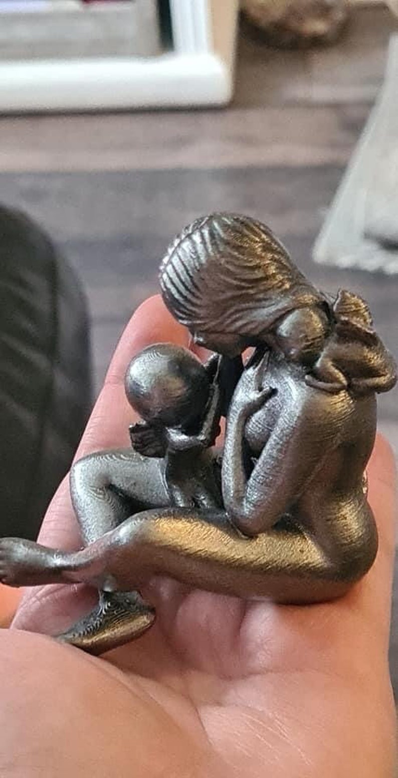 K&ouml;nnte beinhalten: Eine silberfarbene 3D-gedruckte Skulptur einer Mutter und eines Kindes. Die Mutter h&auml;lt das Kind fest an ihrer Brust, und das Kind hat seine Arme um den Hals der Mutter gelegt.