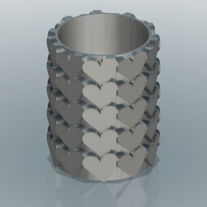 May include: A gray cylindrical container with a pattern of hearts around the outside. The container is made of a smooth, matte material.