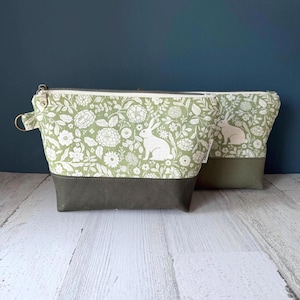 May include: Two zippered pouches. The larger pouch has a light green floral pattern with white rabbits and flowers. The bottom and the smaller pouch are a solid olive green. The pouches are on a white wooden surface with a dark blue background.