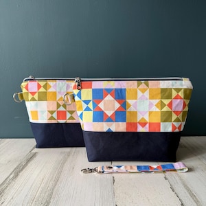 May include: Two zippered cosmetic bags with a colorful patchwork design and a matching wrist strap. The bags have a navy blue base and silver zippers. The patchwork features squares and star patterns in various colors.