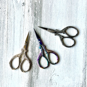 May include: Three pairs of decorative embroidery scissors with ornate handles. One pair is silver, another is iridescent with rainbow hues, and the third is silver. The scissors are on a white and grey wooden surface.