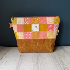May include: A patchwork fabric zipper pouch with a brown waxed canvas base. The top section features a variety of floral and geometric patterned squares in shades of yellow, pink, and peach. A pink zipper and a small metal ring are visible.