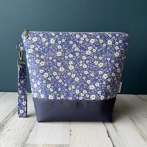 May include: A zipped pouch featuring a floral pattern with white and purple flowers on a blue background. The bottom section is solid navy blue fabric. A matching floral fabric wrist strap is attached to a gold-toned metal clasp on the side.