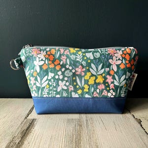 May include: A green zippered pouch with a floral print and a blue bottom. The pouch has a silver zipper and a small tag that says "Sewn with Love".