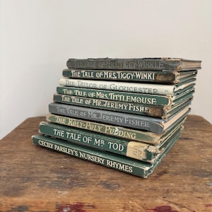 Puede incluir: Una pila de libros infantiles antiguos con cubiertas verdes y blancas. Los títulos incluyen "The Tale of Mrs. Tiggy-Winkle", "The Tailor of Gloucester" y "Cecily Parsley's Nursery Rhymes". Los libros están apilados sobre una superficie de madera.