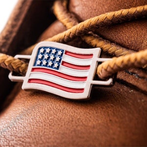 May include: Close-up of a brown leather boot with a silver metal American flag charm attached to the laces.