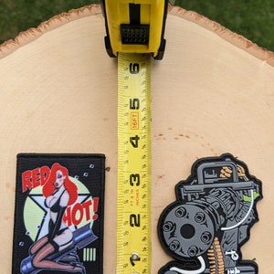 May include: Two embroidered patches, one with a cartoon of a woman in a red dress and the text "RED HOT!" and the other with a detailed illustration of a machine gun. A yellow measuring tape is placed between the patches.
