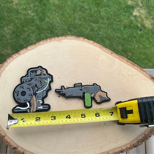 May include: Two black and green military-style patches on a wooden surface. The patches are of a machine gun and a soldier operating a machine gun. A yellow measuring tape is in the foreground.