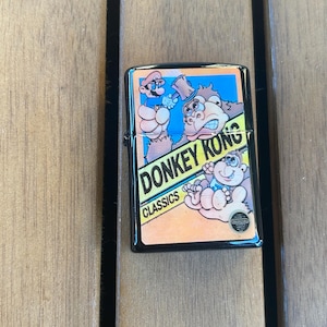 Puede incluir: Encendedor de metal negro con un diseño de Donkey Kong Classics. La obra de arte incluye a Mario, Donkey Kong y un bebé Kong, sobre un fondo amarillo y azul. El encendedor es rectangular y tiene un acabado brillante.