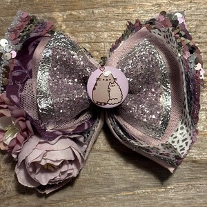 Puede incluir: Un lazo de purpurina morado y plateado con un adorno floral y un botón con un gato de dibujos animados con un corazón.