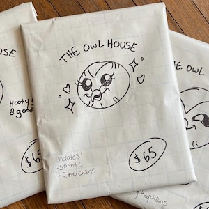 May include: Three white packages with hand-drawn owl illustrations and the text "THE OWL HOUSE." Each package has a price tag of $65 and includes prints and keychains. The packages are on a wooden surface.